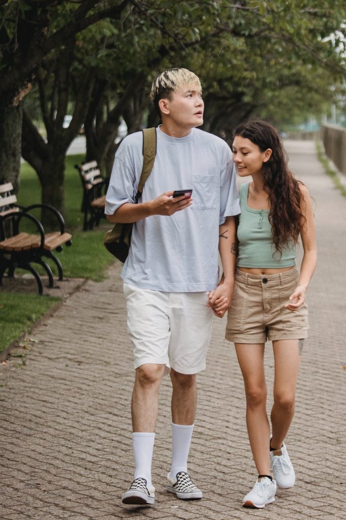 loving ethnic couple walking along park alley and using smartphone