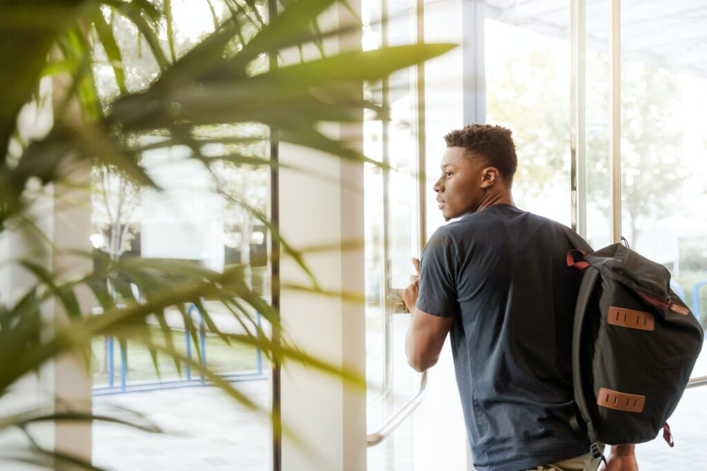 man looking outside window carrying black and brown backpack while holding his hand on window